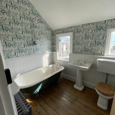 A bathroom scene with a claw footed tub, a sink and a loo. The bathroom walls have been wallpapered using Louise Altman's hand block printed Lucy design in a dark green pattern on a light warm white ground.