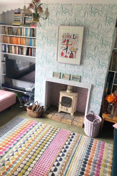 A high up view point looking don onto a lounge. The lounge has a wood burning stove and a richly patterned rug. The main focus of the image is the hand block printed wallpaoer which adorns the walls. the colour of the wallpaper is a pale grey with a green teal coloured leaf pattern.
